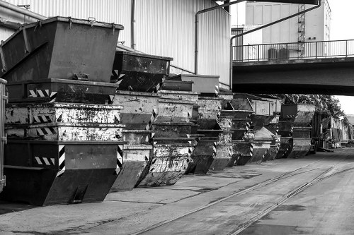 A row of large metal skips filled with mixed waste materials, positioned along a concrete driveway adjacent to a warehouse or industrial building. The skips are weathered, with visible rust and peeling paint, and are stacked in a straight line stretching from the foreground to the background. The surface of the skips shows scratches and dents, indicating frequent use in waste management operations. Overhead, a small portion of an elevated walkway or platform is visible, along with some piping or conduit attached to the building's exterior. The environment appears to be an industrial or commercial area, possibly in Bayswater, where Business Waste Removal Bayswater offers rubbish removal services. The lighting seems natural, with soft shadows suggesting an outdoor setting in daytime, and the overall scene reflects a professional waste collection or disposal site appropriately related to rubbish removal services.