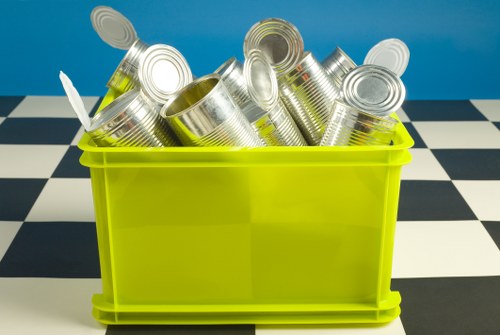 A bright lime-green plastic storage container filled with numerous aluminium cans, some with visible ridges and pull-tab tops, placed on a black and white checkered floor against a solid turquoise background. The cans are stacked haphazardly, with some leaning against each other and a few with open tops revealing their smooth, silver interior. The container appears to be a waste collection bin used for rubbish or recyclable materials, commonly found in waste removal settings in Bayswater or nearby areas. The scene is well-lit, highlighting the metallic sheen of the cans and the vibrant color of the bin, emphasizing the typical environment for rubbish disposal or recycling preparation within a domestic or commercial premises serviced by Business Waste Removal Bayswater or similar waste management companies.