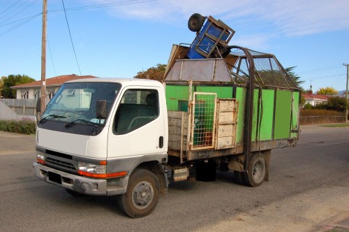 A small flatbed truck used for rubbish removal, with a white cab and a wooden cargo area featuring green panels and metal fencing on the sides. The cargo area is loaded with various discarded items, including a blue plastic chair positioned at an angle on top of a pile of mixed waste materials. The vehicle is parked on a paved street in a residential area, with houses, trees, and a utility pole visible in the background under a clear blue sky. The truck appears weathered, indicating it has been used regularly for waste collection or clearance services. This image illustrates typical rubbish removal equipment deployed by a local waste management service, such as Business Waste Removal Bayswater, operating within the Bayswater postcode area and surroundings. The scene captures the vehicle in daytime light, emphasizing the textures of the wooden and metal surfaces, as well as the casual stacking of waste items for collection. The presence of waste materials and the type of vehicle suggest an example of a domestic or small-scale commercial rubbish collection operation.