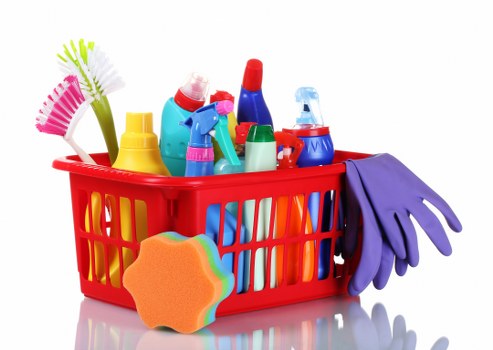 A red plastic basket filled with various cleaning supplies and tools, including spray bottles, a scrub brush, rubber gloves, and a multi-colored sponge, positioned on a reflective surface. The cleaning products are made of plastic with bright colors such as blue, yellow, red, and green. The rubber gloves are purple and draped over the side of the basket, while the sponge features a multicolored, rainbow-like edge. The background is white with even lighting, emphasizing the neatly arranged items commonly associated with domestic or commercial cleaning tasks, relevant to waste removal services like those offered by Business Waste Removal Bayswater in the Bayswater area, suggesting a focus on waste collection of cleaning supplies or similar items.
