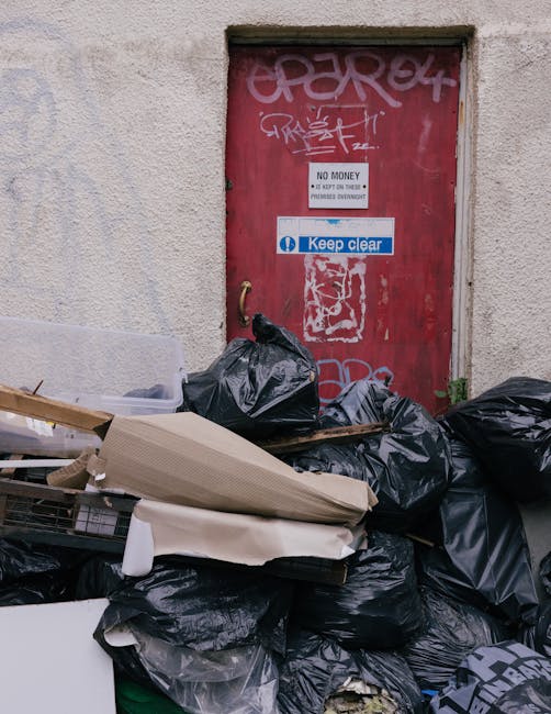A collection of black garbage bags filled with waste piled against a textured, off-white concrete wall beneath a small, rectangular, red painted metal door that has graffiti tags in white and pink spr