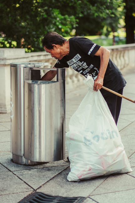 A collection of black garbage bags filled with waste piled against a textured, off-white concrete wall beneath a small, rectangular, red painted metal door that has graffiti tags in white and pink spr