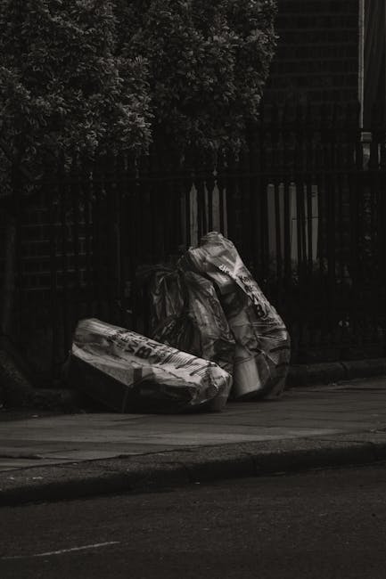 Two large black plastic garbage bags filled with refuse are placed on a dark pavement beside a curb, leaning against a black metal fence with vertical bars. The bags appear to contain household waste 