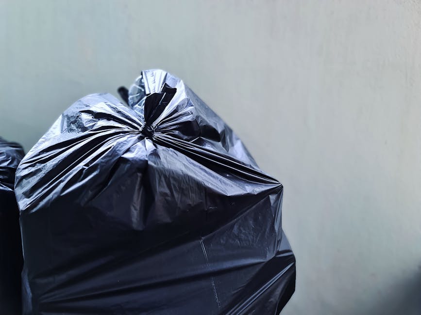 Two large black plastic garbage bags filled with refuse are placed on a dark pavement beside a curb, leaning against a black metal fence with vertical bars. The bags appear to contain household waste 